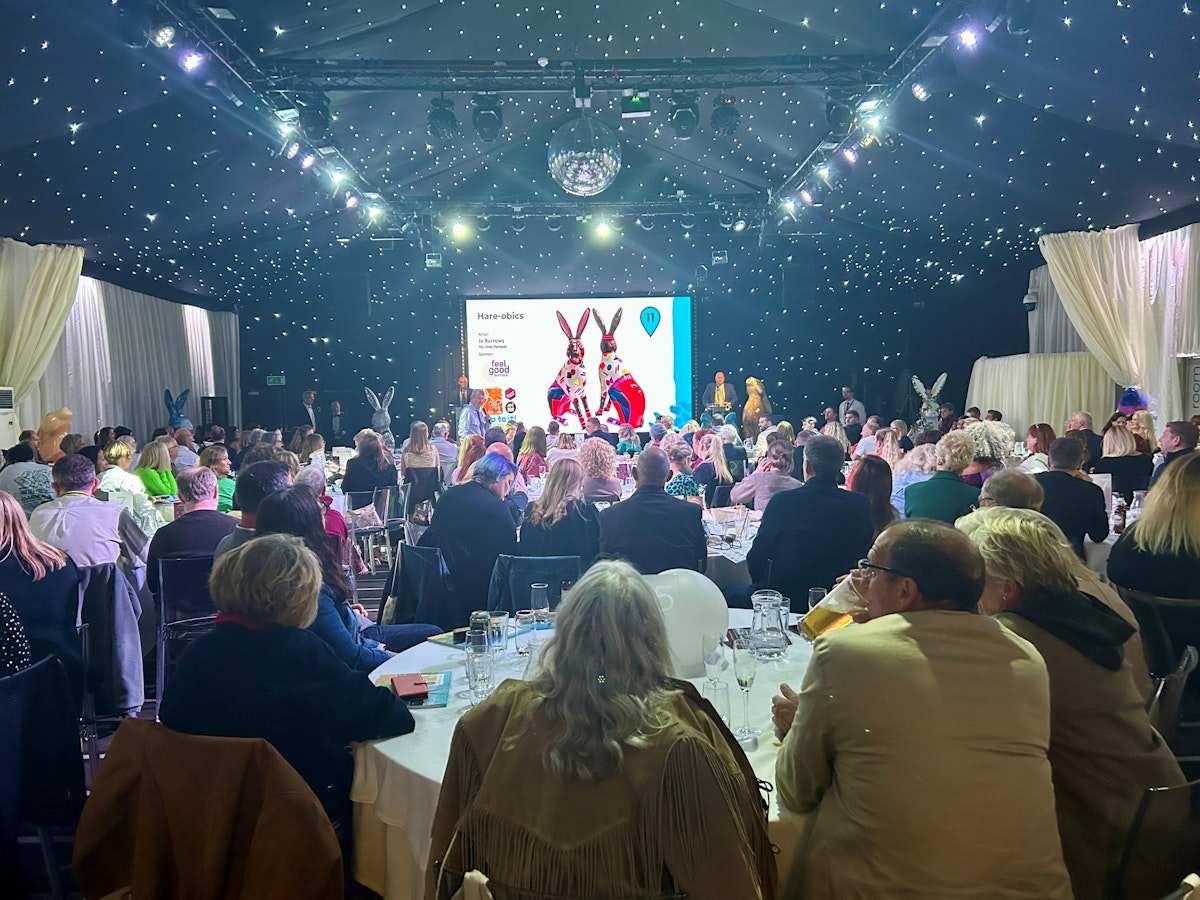 Large crowd seated at tables watching the stage during the hospice hare auction, with sculptures visible in the background